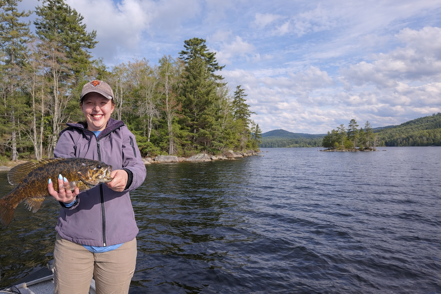 Angler holding a smallmouth bass on a Maine lake, standing on a boat with forested shoreline and mountains in the background.