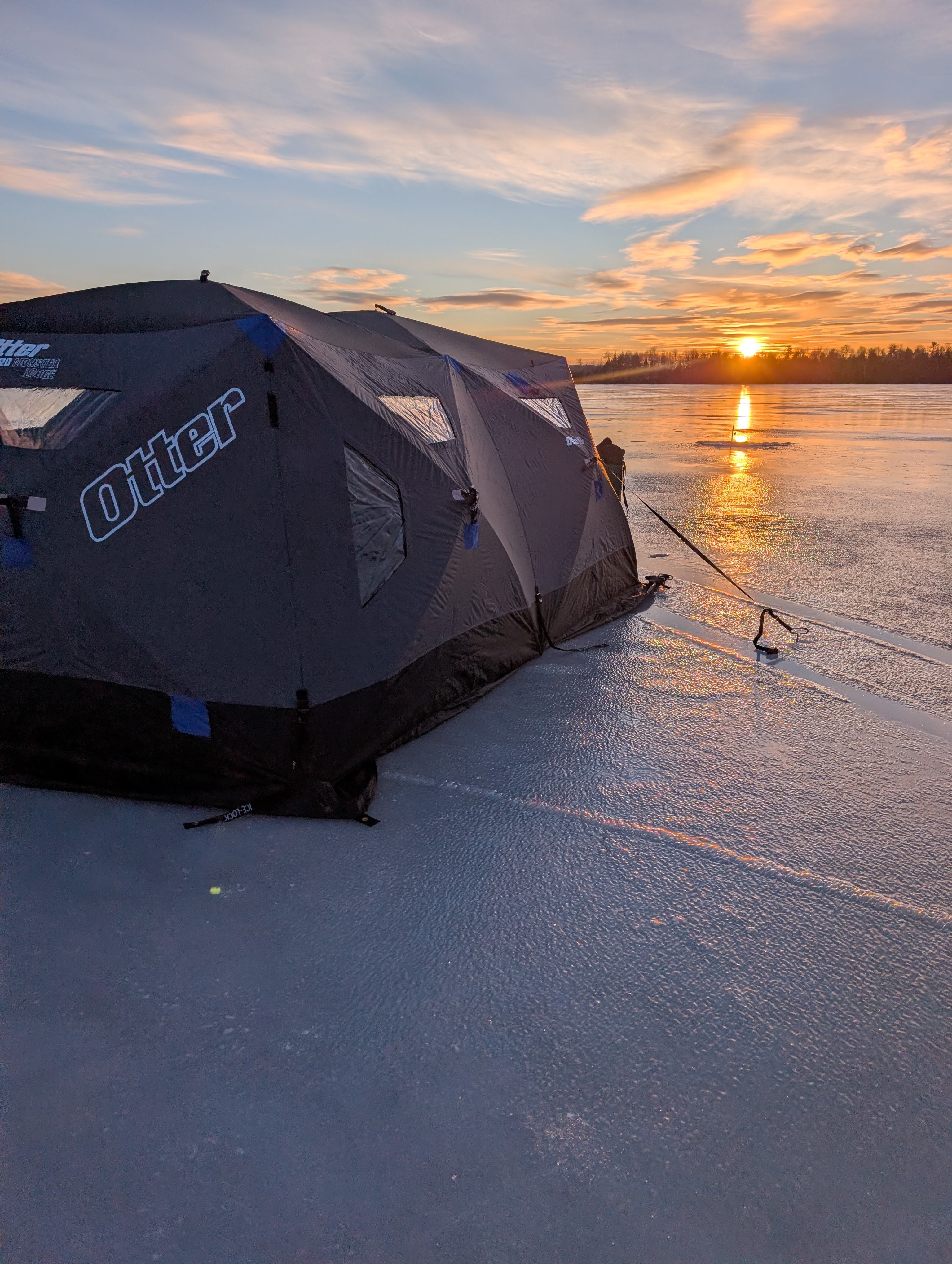 Otter ice fishing shelter set up on a frozen lake in Maine at sunset with golden light reflecting across the ice.