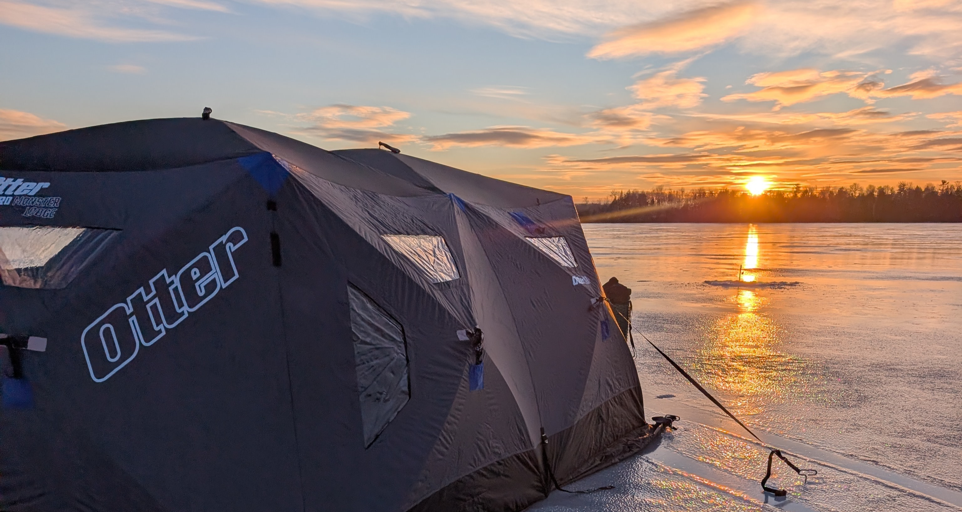 Otter ice fishing shelter set up on a frozen lake in Maine at sunset with golden light reflecting across the ice.
